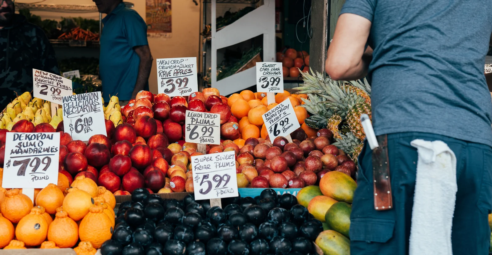 Puesto de mercado con letreros de precios escritos a mano para distintas frutas, ejemplo de variación de precios por producto y punto de venta